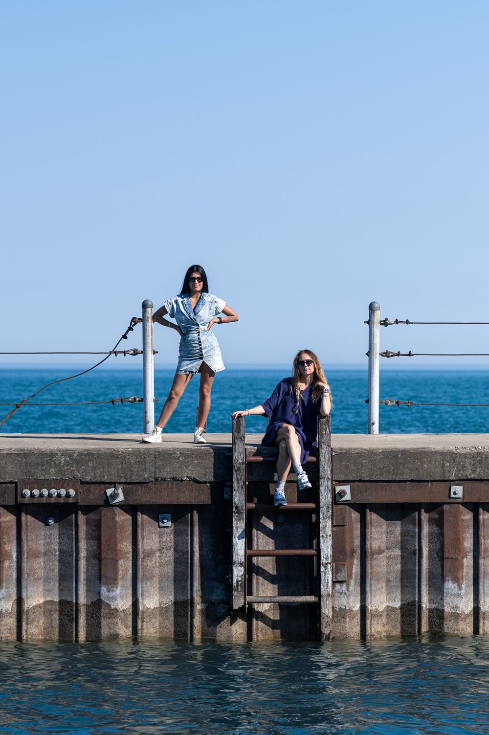 Two people posing on a pier railing with a blue body of water and sky backdrop.