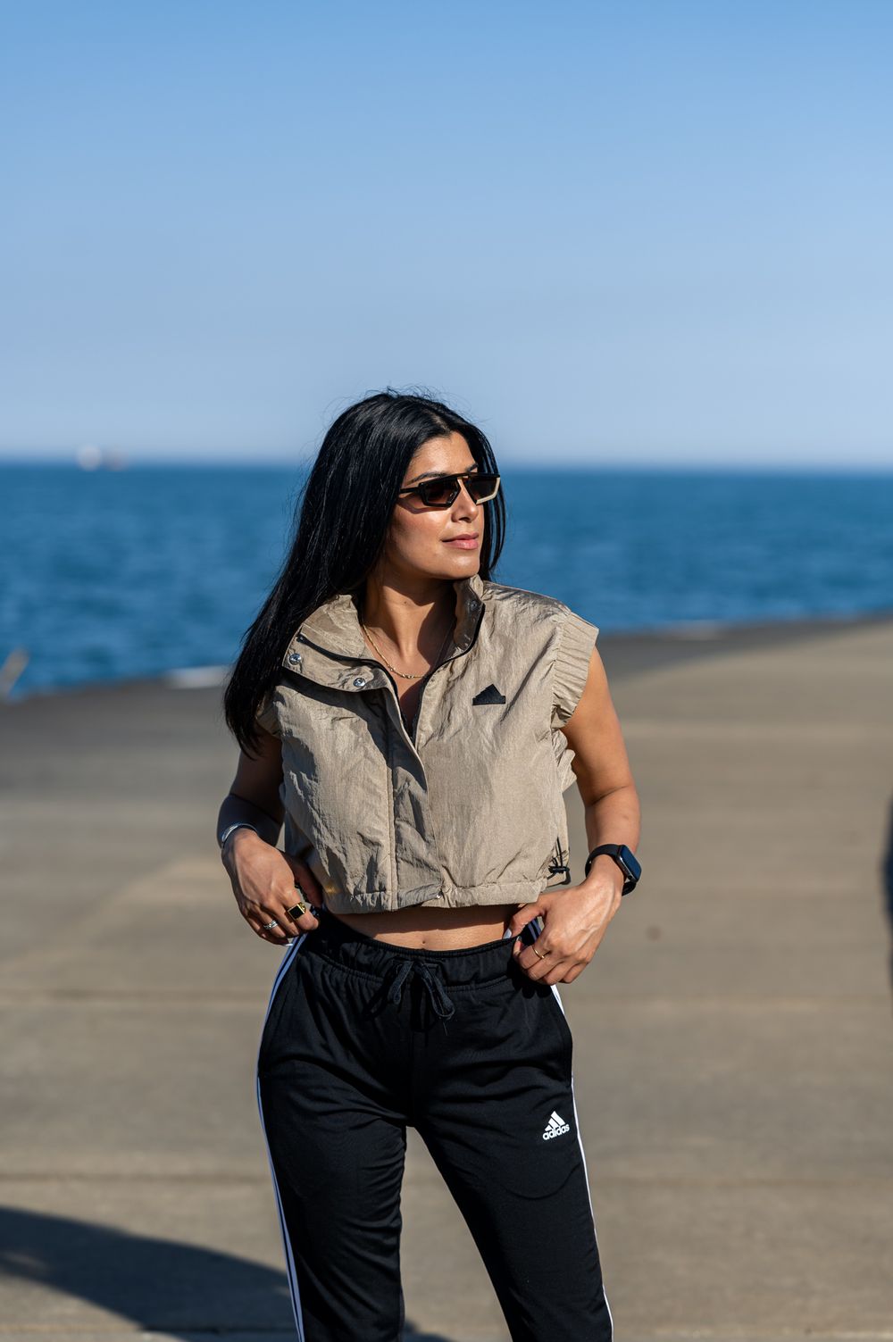 Stylish casual outfit featuring a beige cropped vest and black Adidas pants against a body of water backdrop.