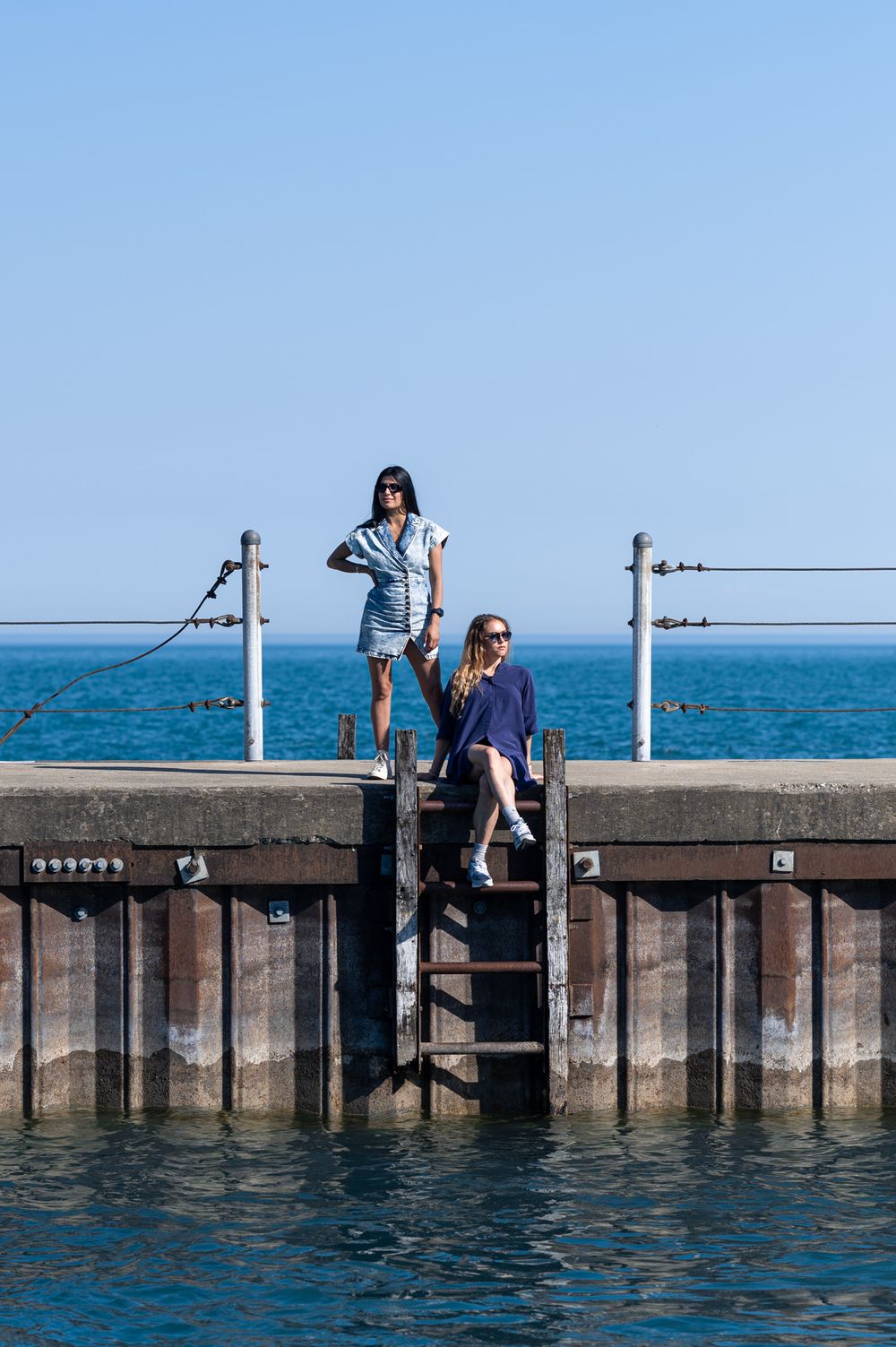 Two people standing on seaside pier stairs against bright blue water front background.