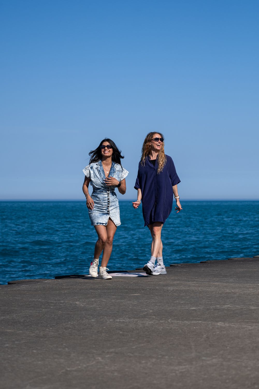 Two friends walking together on oceanfront pier in casual summer outfits.