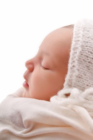 Peaceful newborn baby sleeping in a white knit bonnet hat, wrapped in soft white blanket, profile view.