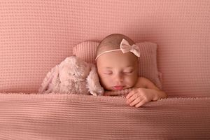 Newborn baby girl sleeping peacefully on pink blanket with stuffed bunny and bow headband in soft pink studio setup.