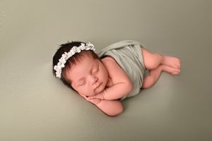 Sleeping newborn baby girl wrapped in sage green swaddle with white floral headband on matching green background.