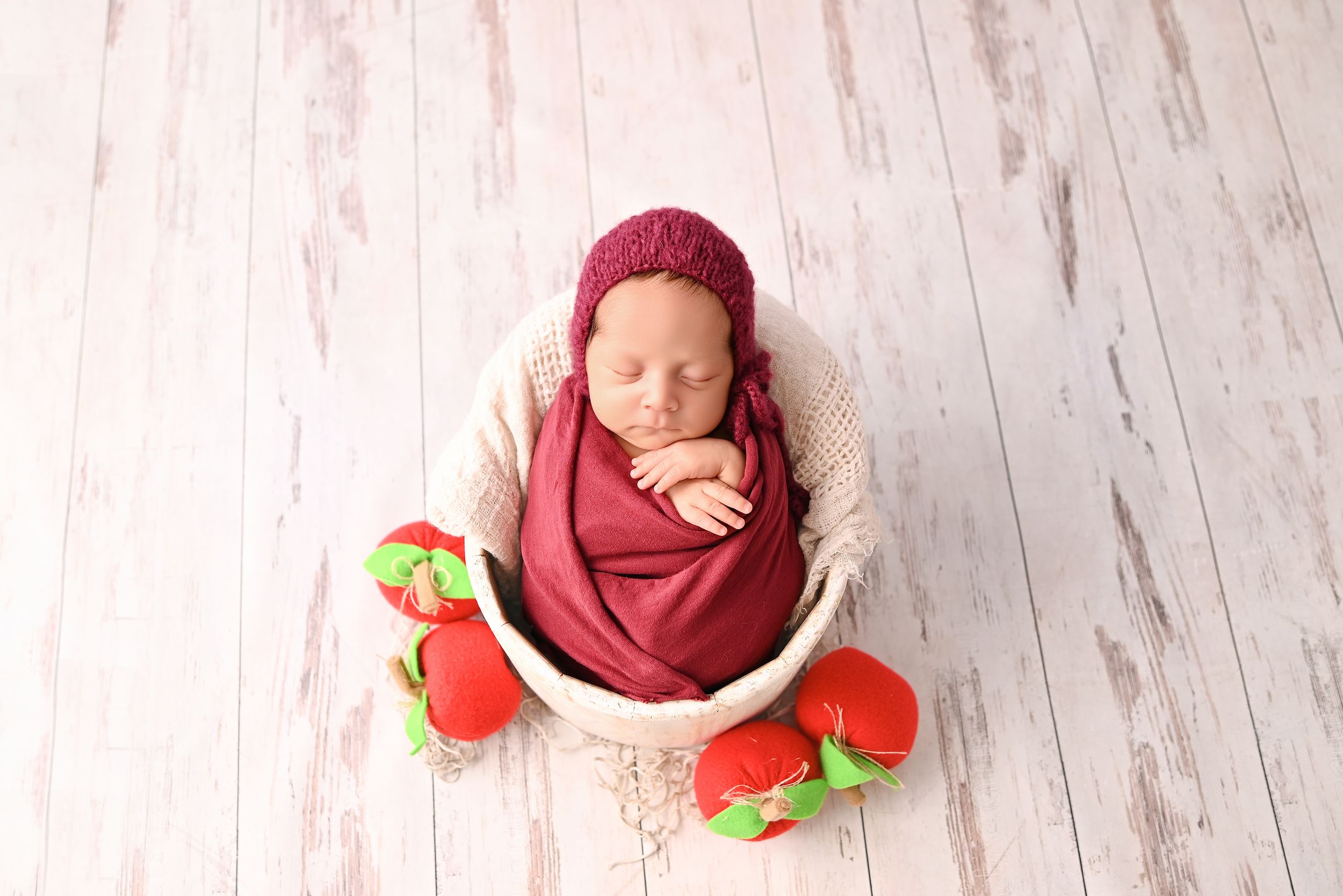 Newborn baby wrapped in burgundy swaddle with matching bonnet, posed in white basket with red strawberry booties.