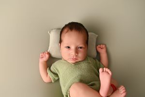Newborn baby in green onesie lying on sage green backdrop with small pillow, arms raised, looking at camera.