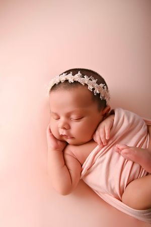 Sleeping newborn baby girl wearing a floral headband, wrapped in a soft pink blanket on a pink background.