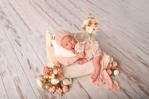 Newborn baby girl sleeping peacefully in a tiny white bed surrounded by soft pink flowers and wearing a delicate pink outfit.