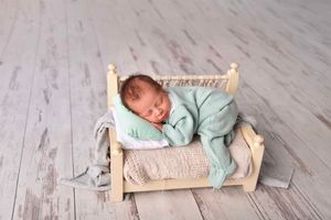 Baby in mint green sleeper crawls across light wooden floor while pulling small wooden toy bed behind them.