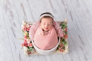 Newborn baby swaddled in pink fabric sleeping in white bowl surrounded by pink and white flowers.
