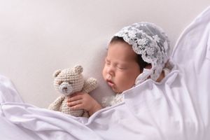 Sleeping newborn with lace bonnet holding teddy bear on white bedding.