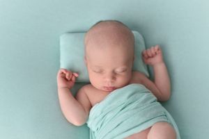 Sleeping newborn baby wrapped in soft mint green swaddle on simple backdrop.