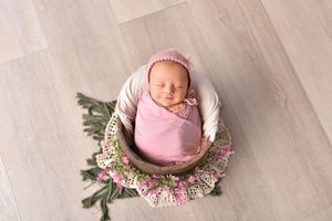 Newborn baby in pink wrap lying on wooden floor surrounded by pink and white flowers and eucalyptus leaves.