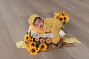 Newborn in yellow outfit sleeping among bright sunflowers on wooden floor.
