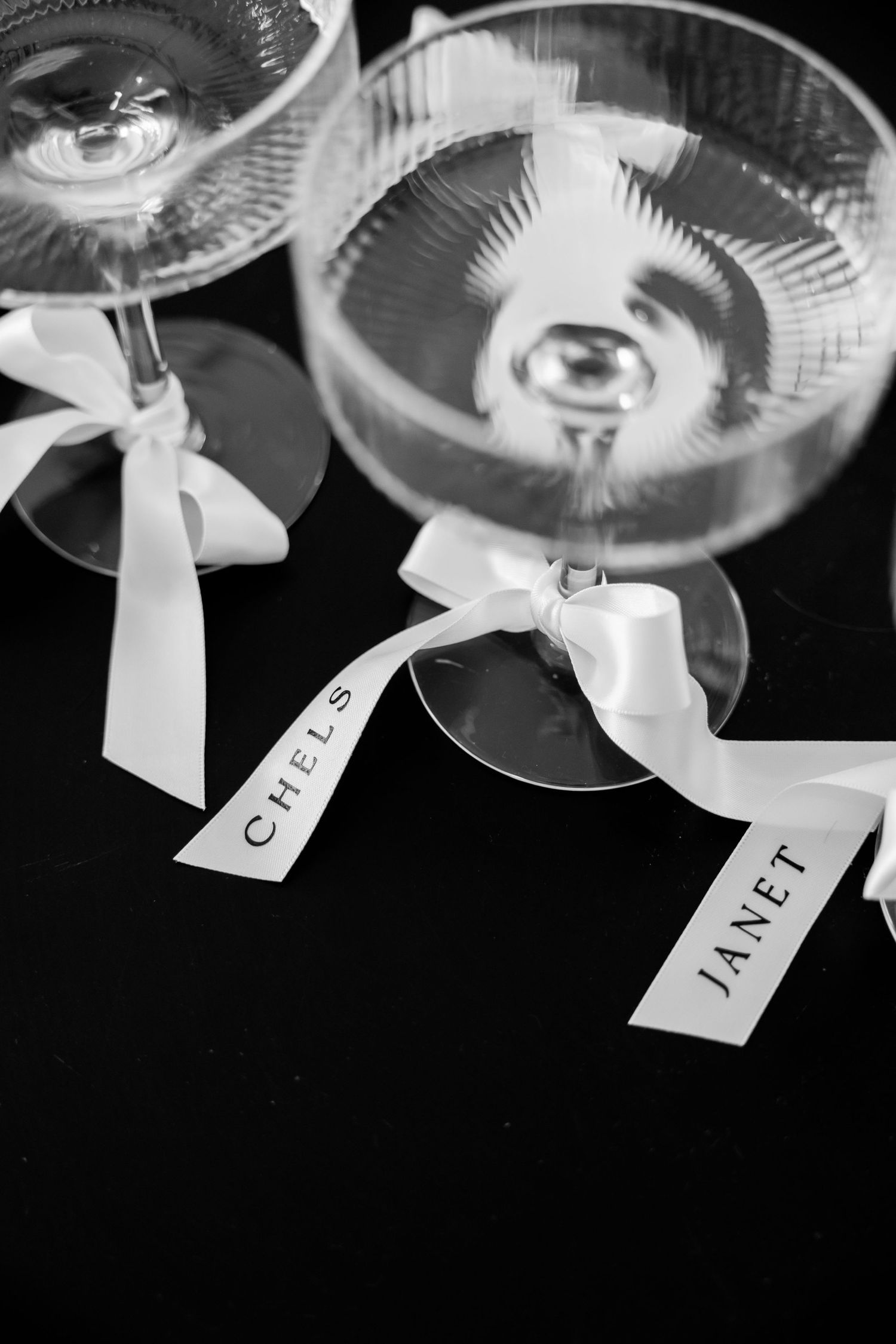 Two martini glasses with white ribbon bows and name tags against a black background in black and white photography.