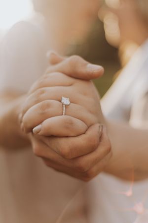 Close-up of clasped hands showing off a sparkling engagement ring in warm sunlight.