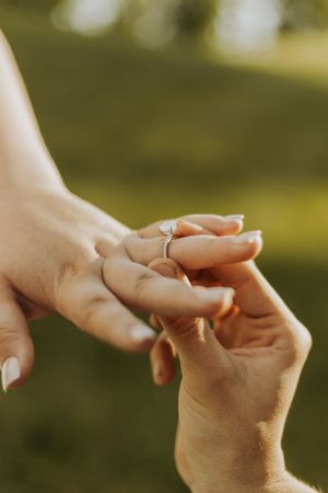 A close-up shot of someone placing an engagement ring on another person's finger outdoors.