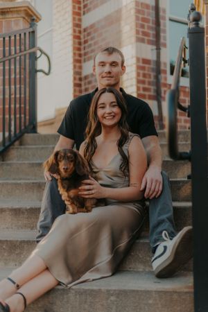 A couple sits on concrete steps with their dachshund puppy in an urban setting.