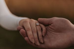 Two hands gently clasped together showing off an engagement ring in warm lighting.