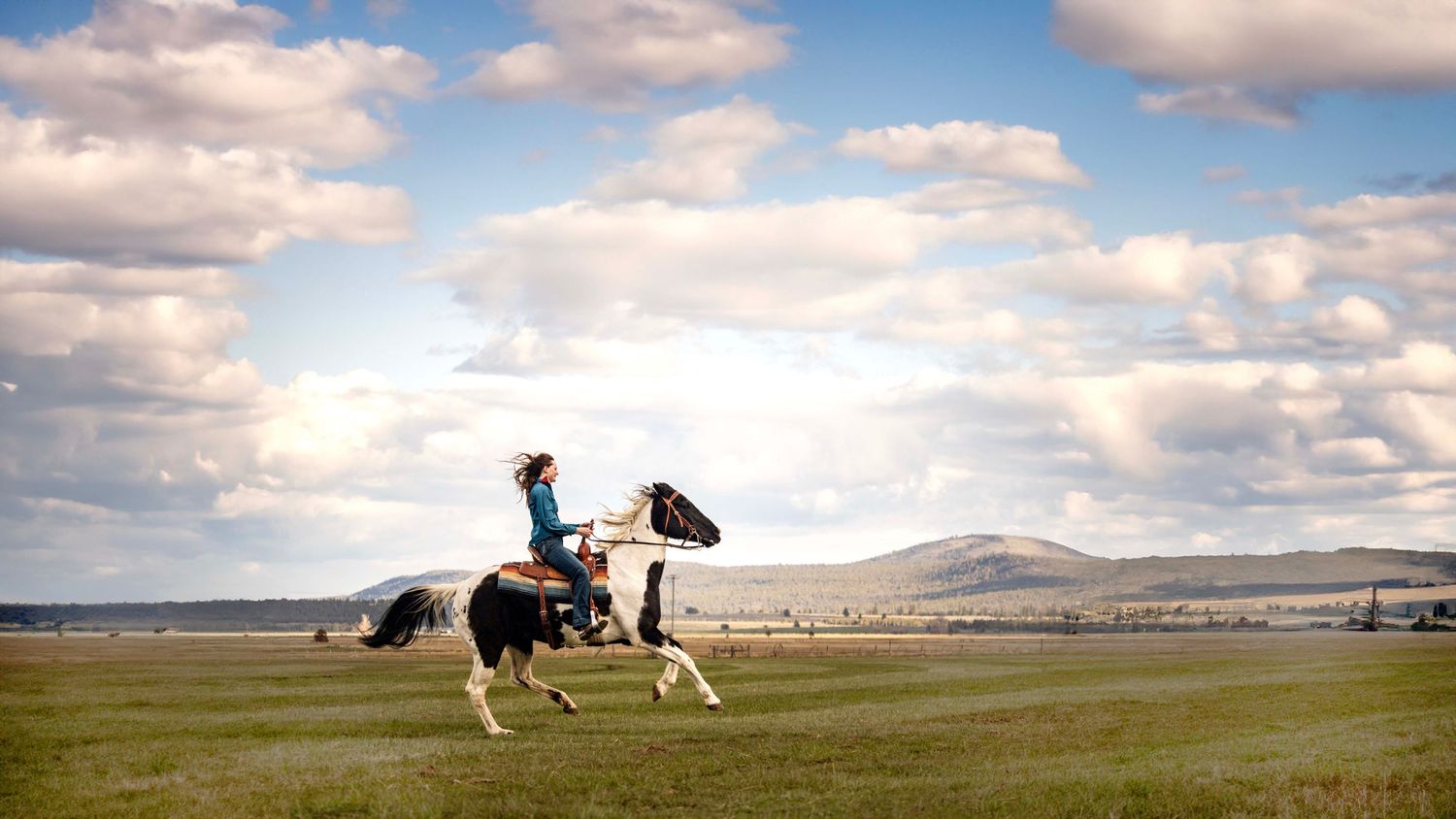 girl dressed in blue rides black and white horse through field as hills loom in distance and the cloudy sky opens up above them