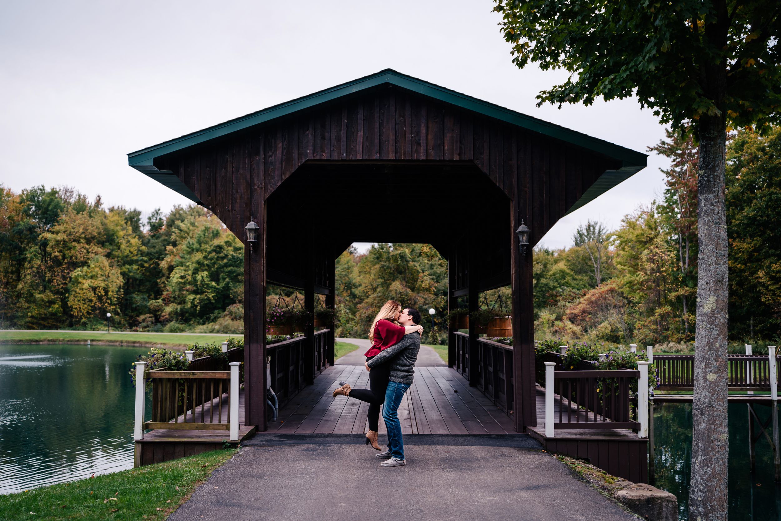 A couple embraces on a wooden covered bridge over a serene lake surrounded by autumn foliage.