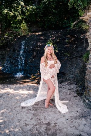A maternity photo of a woman in lace dress posing by a waterfall.