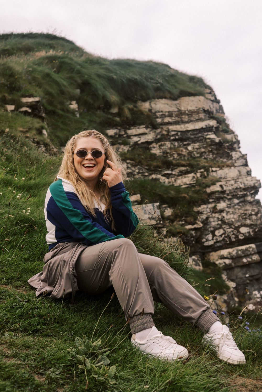 Person in sunglasses and athletic wear sits on grassy cliffside overlooking scenic coastal landscape in Ireland.