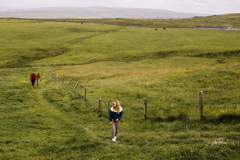 Two people walk along a winding fenced path through green rolling hills near the coast on a cloudy day.