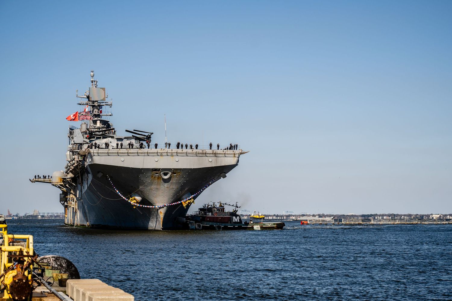 Aircraft carrier docked in harbor under clear blue sky with crew visible on deck.