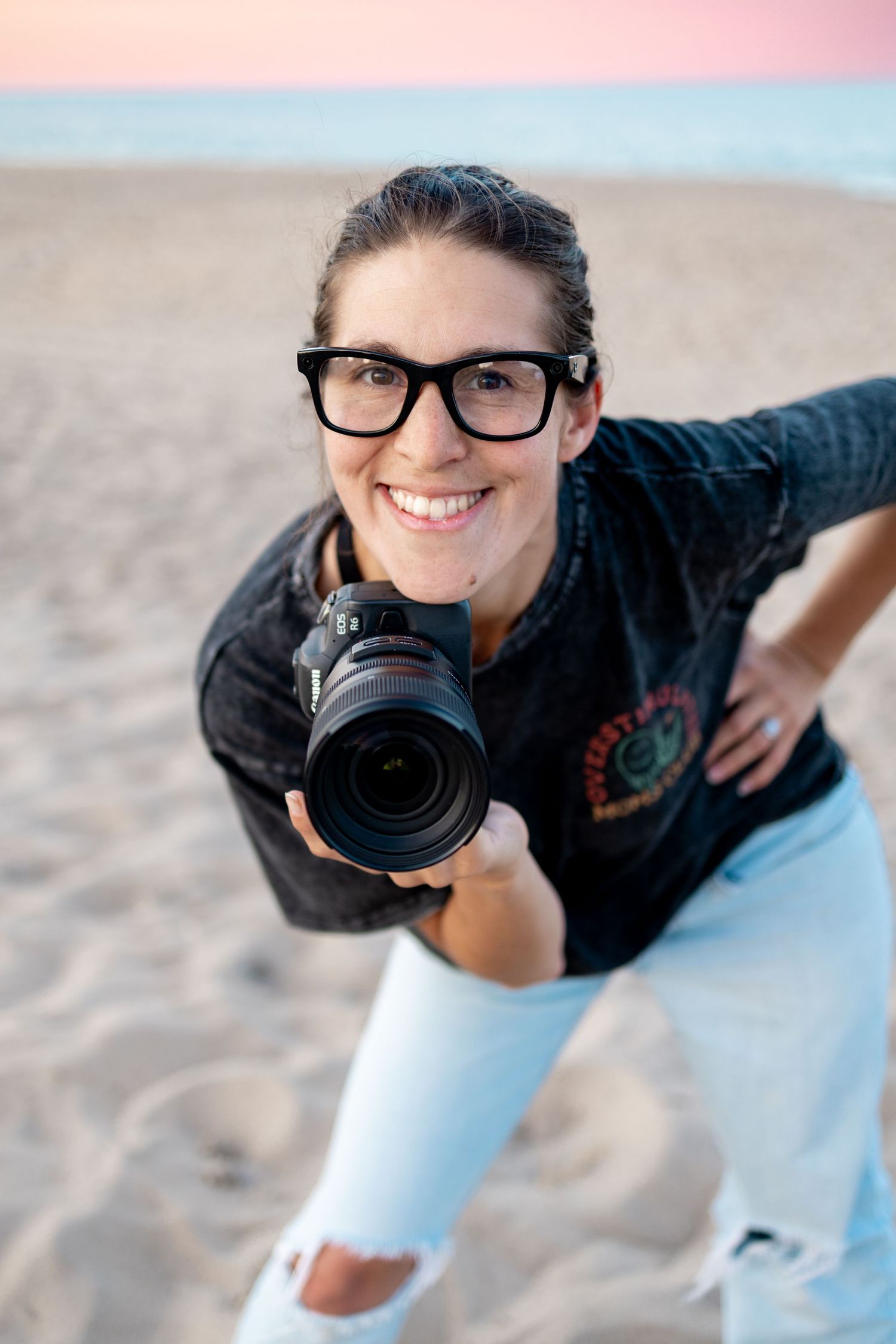 Woman photographer smiling on beach, holding DSLR camera, wearing black glasses and denim jacket, with sandy shore background.