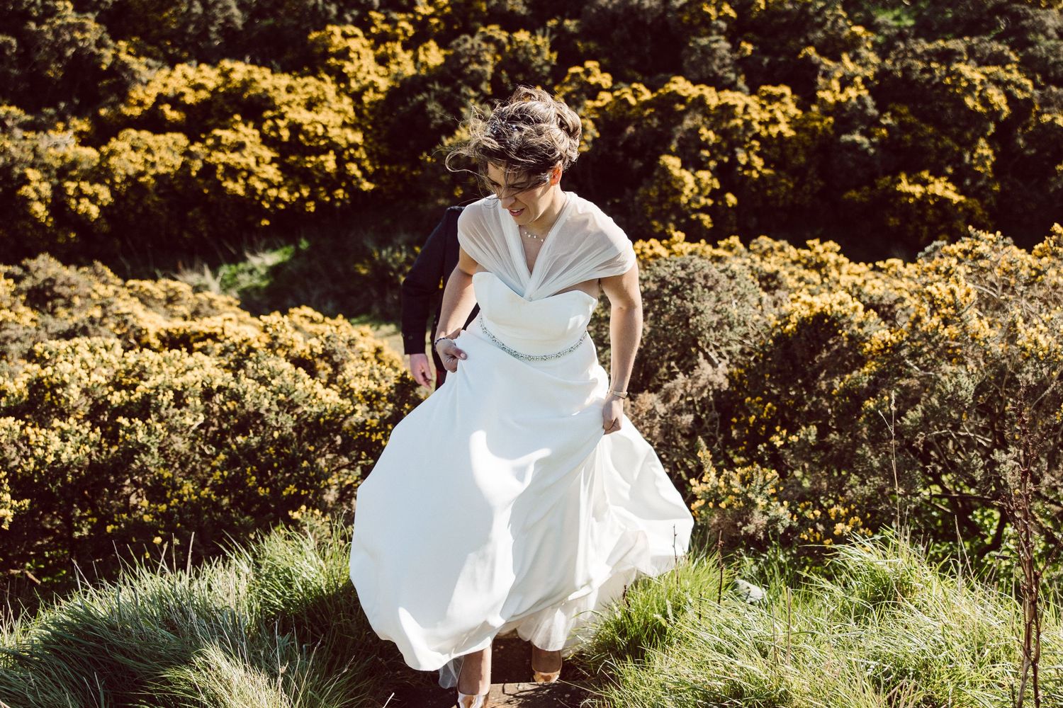A couple in wedding attire walking through blooming yellow gorse on a hillside.