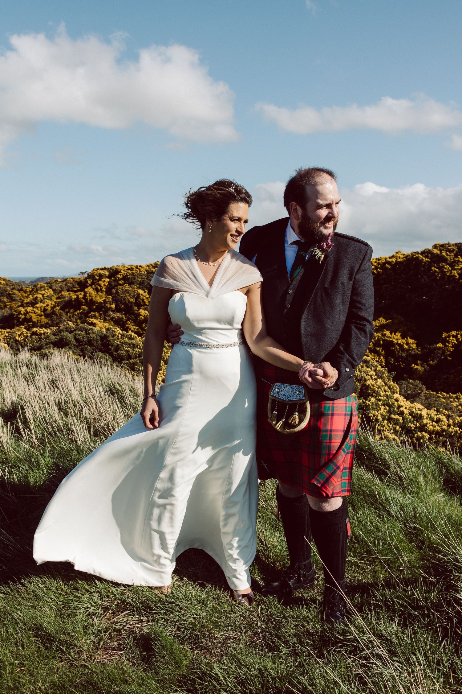 Couple in wedding attire and Scottish kilt walking through tall grass and yellow gorse.