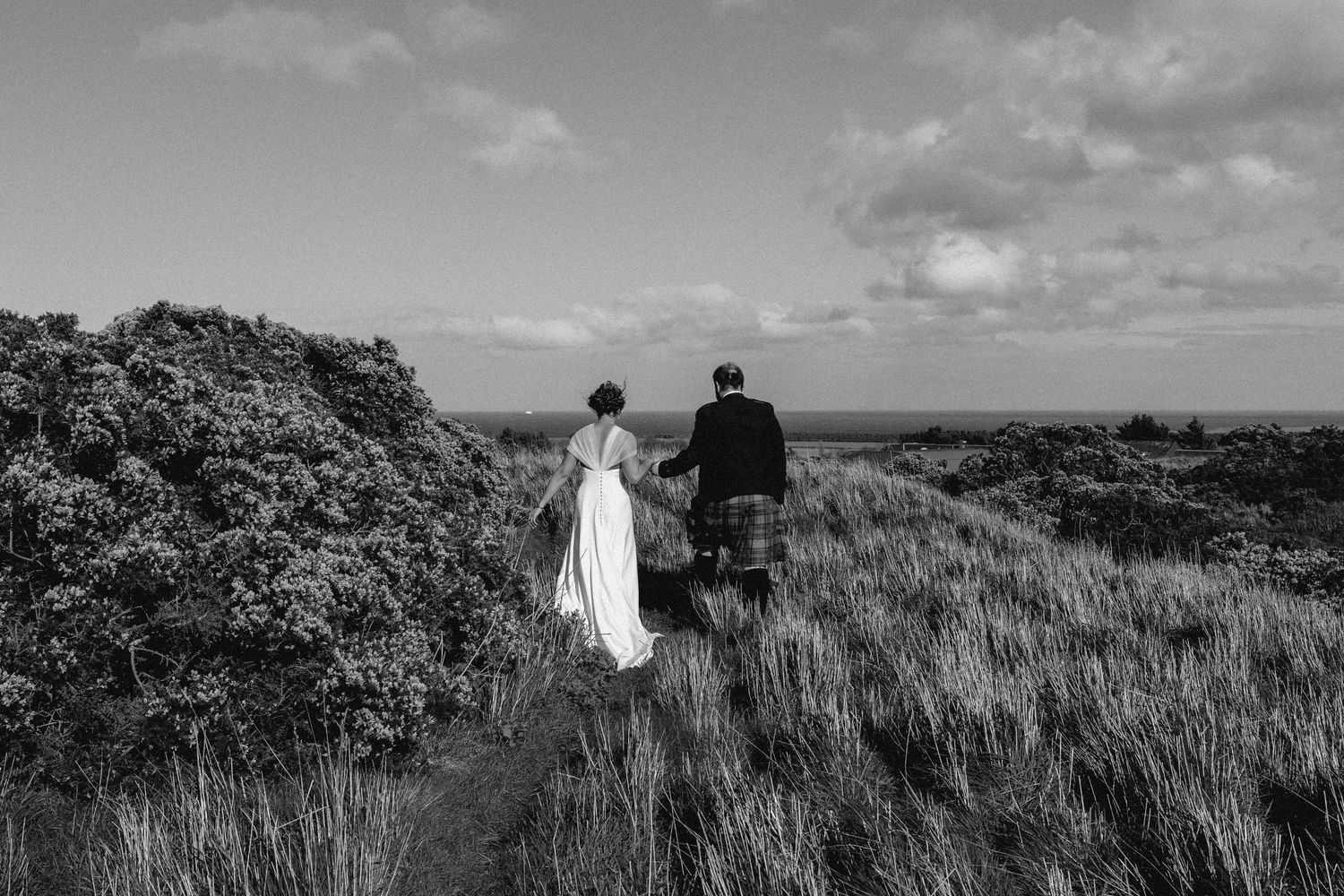 Black and white photo of a couple walking through grassy hillside with ocean view.
