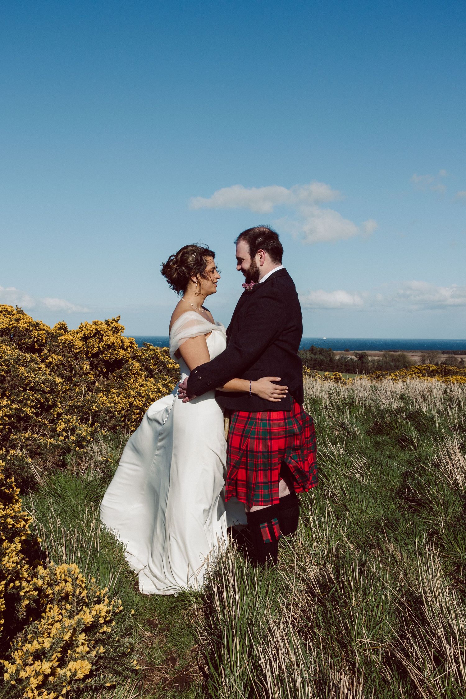 Wedding couple embracing on coastal hillside surrounded by yellow gorse and ocean views.