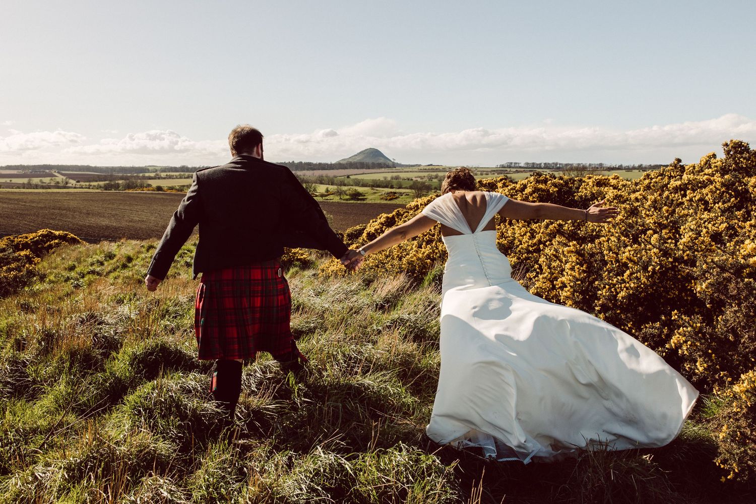 A couple in wedding attire walks hand in hand across a scenic hilltop with mountains in the distance.