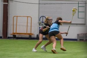 Two lacrosse players battle for position during an indoor practice session on artificial turf.