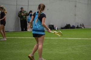 Lacrosse player in light blue jersey holding stick while moving across indoor field.