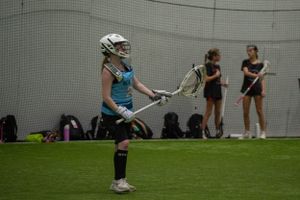 A player practices lacrosse stick handling while others observe in indoor facility.