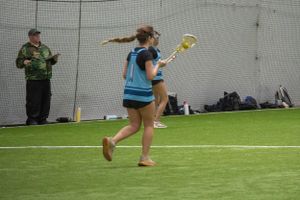 A player runs with a yellow lacrosse stick during indoor practice as coach observes.