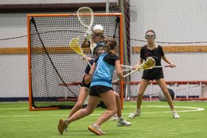 Players engage in competitive lacrosse drill near the goal during indoor practice.