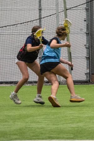 Two players compete for position during an intense indoor lacrosse practice drill.