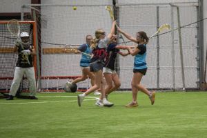 Players in black and blue uniforms compete intensely during an indoor lacrosse match on artificial turf.