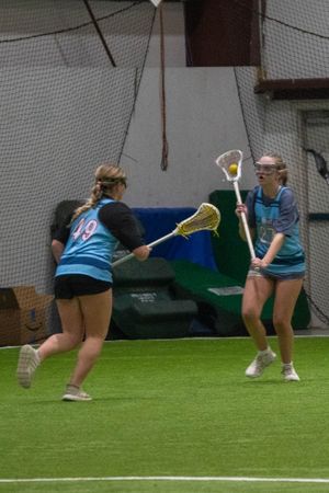 Two players practice lacrosse passing drills on an indoor turf field with protective netting.