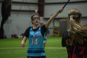 Indoor lacrosse match showing players competing with sticks raised.