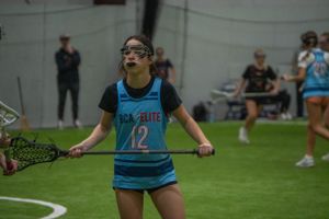 A player in blue jersey displays intense concentration during an indoor lacrosse game.