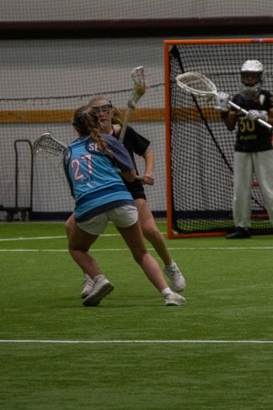 A player takes a dynamic shooting stance near the lacrosse goal with a goalie defending.