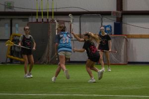 Indoor lacrosse players competing in a practice match with players in blue and black uniforms on artificial turf.
