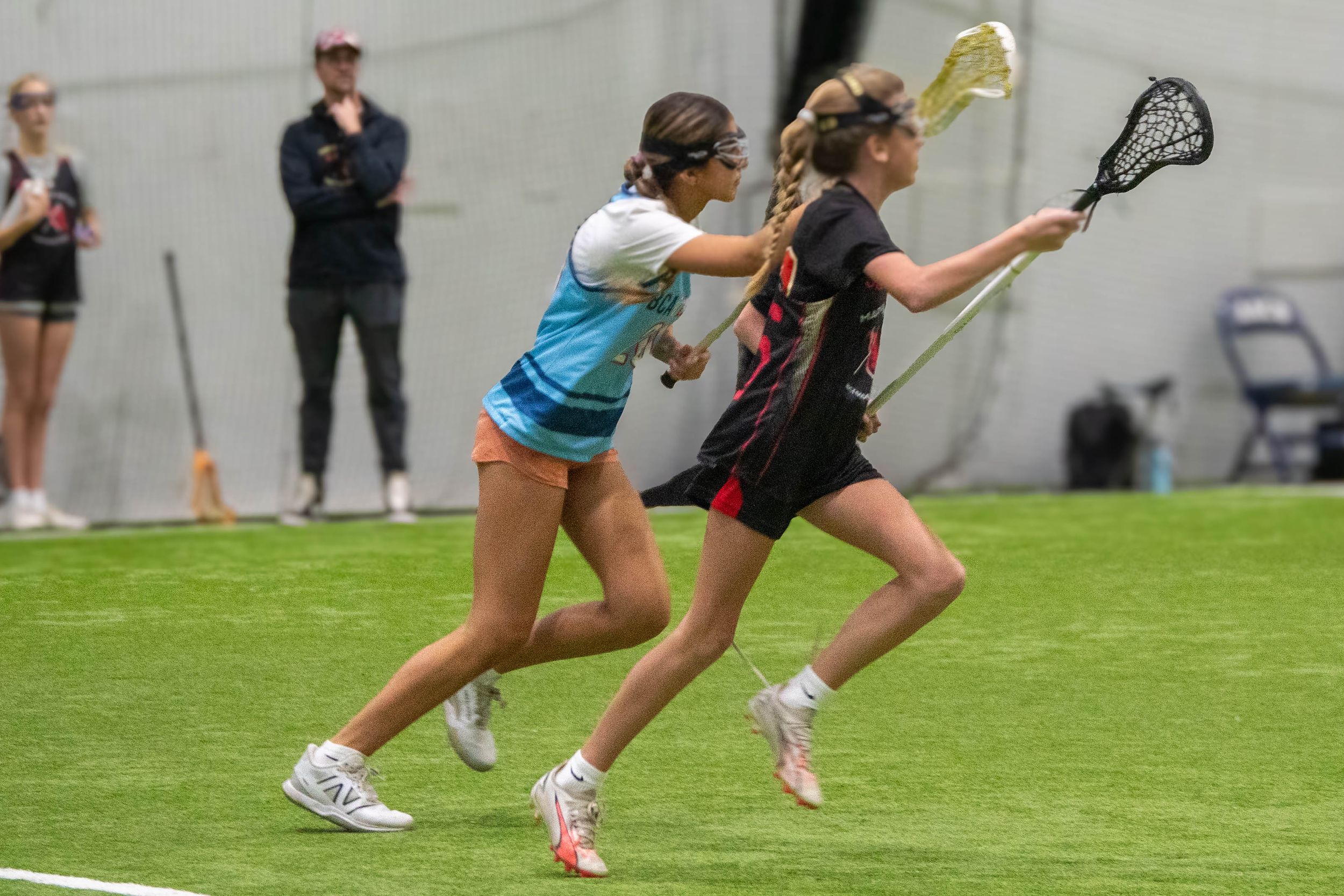 Two players competing in an indoor lacrosse match on artificial turf.