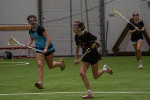 Athletes in black and blue uniforms running with lacrosse sticks during an indoor practice session.