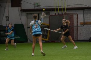 Players compete for control of a yellow ball during an indoor lacrosse match.