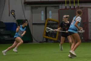 Indoor lacrosse players maneuvering with sticks during an intense gameplay moment.